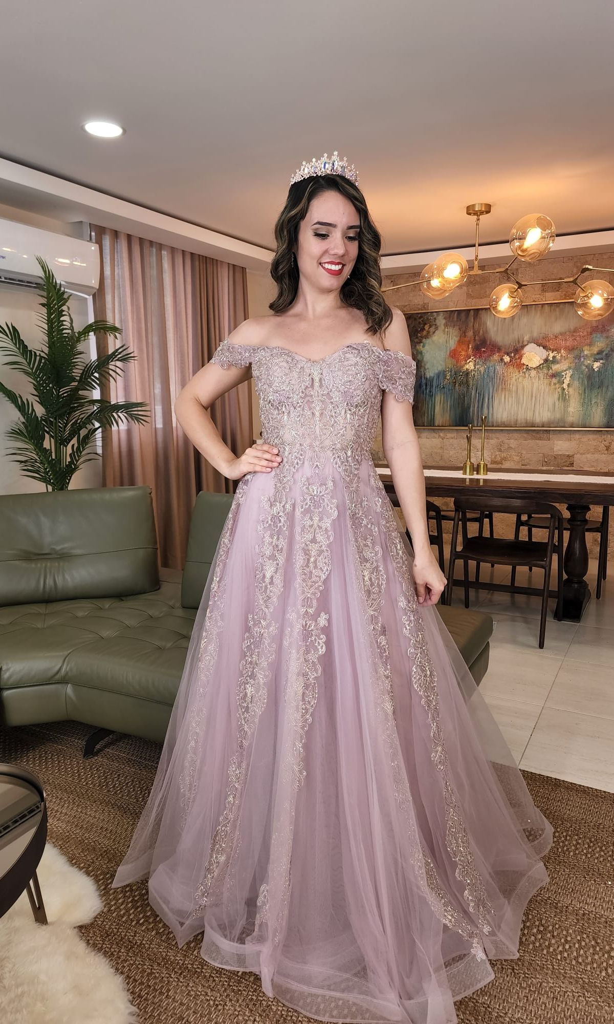 A woman in the Chicas CA1103 Long Lace Prom Ball Gown with off-shoulder design and intricate embroidery stands in a modern living room with green furniture, stylish wall art, and a tiara on her head.